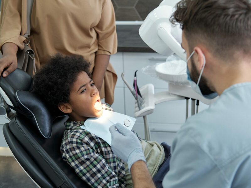 Youthful African boy with open mouth looking at male dentist with instruments before oral check-up
